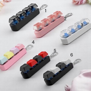 May include: Six keychains with mechanical keyboard keycaps in different colours. Keycaps are black, pink, white, red, and blue. Each keychain has a silver loop for attachment. The keycaps are made of plastic.