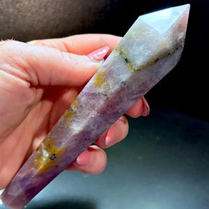 May include: A polished, hexagonal crystal point featuring a blend of translucent lavender and opaque white mineral sections. The stone displays natural inclusions of golden-yellow and dark grey flecks throughout its elongated, tapered structure.