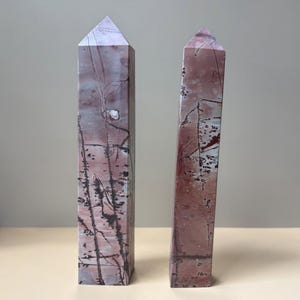 May include: Two tall, polished stone towers with pointed pyramid tops. The stones feature a muted red and dusty pink base color with intricate, dark brown dendritic patterns and thin, branching lines running vertically across the surfaces.