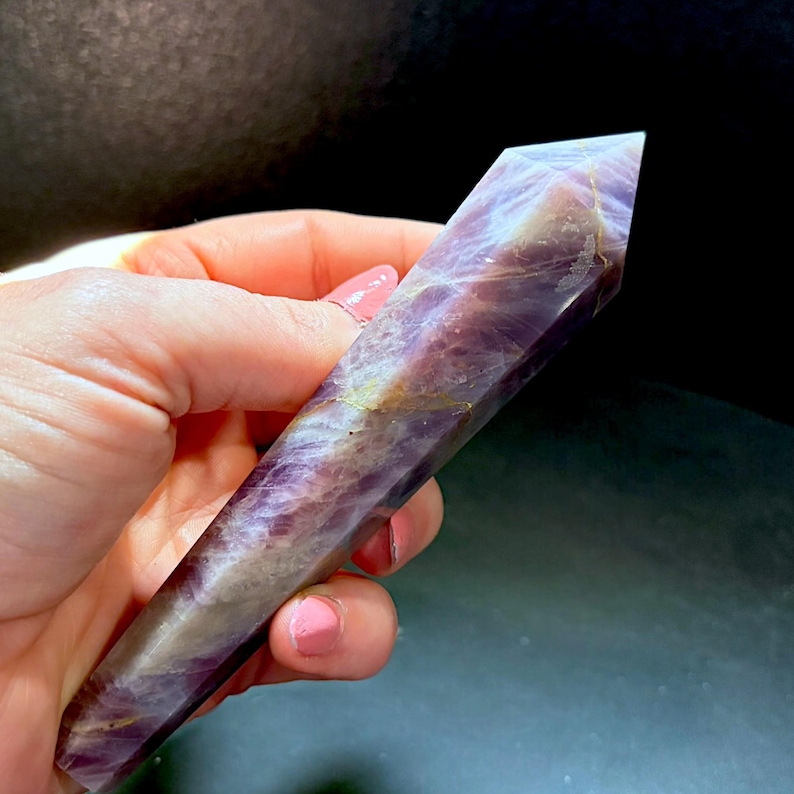 May include: A polished, hexagonal crystal point featuring a blend of deep purple amethyst and translucent white quartz. The stone displays natural internal fractures, cloudy inclusions, and subtle golden-brown mineral veins throughout its length.