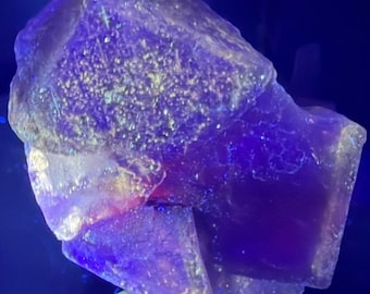 May include: A raw, crystalline mineral specimen featuring deep purple and violet hues. The surface displays a jagged, multifaceted texture with translucent sections that catch the light, creating a glowing effect under intense blue illumination.