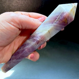 May include: A polished, six-sided crystal tower featuring a blend of translucent white and deep purple hues. The stone exhibits natural internal veining and a pointed termination, showcasing a smooth, faceted surface with varied color saturation.