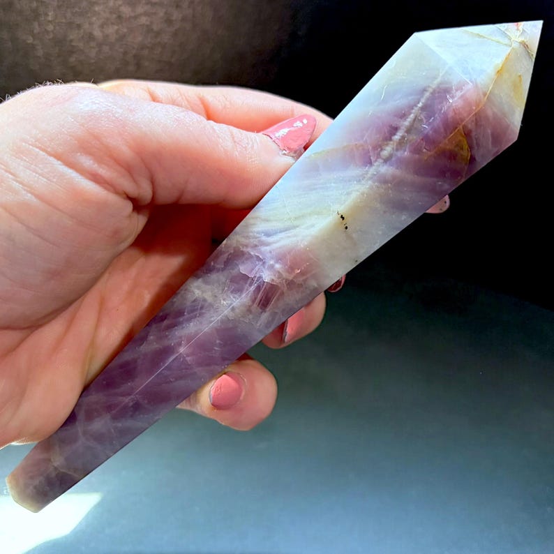 May include: A polished, six-sided crystal tower featuring a blend of translucent white and deep purple hues. The stone exhibits natural internal veining and a pointed termination, showcasing a smooth, faceted surface with varied color saturation.