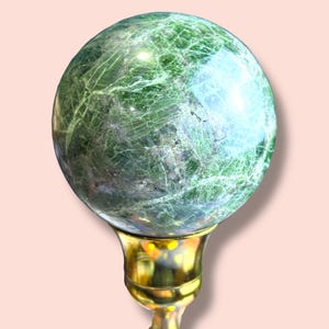 May include: A green sphere with a marbled appearance, mounted on a gold-colored base. The sphere has a polished surface with light and dark green veining. The base has a decorative, ornate design.