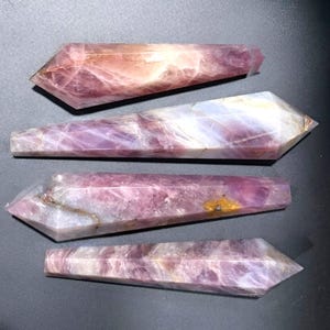 May include: Four polished crystal wands arranged horizontally. Each wand features a faceted point and a hexagonal body with varying shades of translucent purple, lavender, and white, with some pieces showing internal veining and small inclusions.