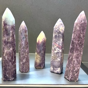 May include: Five polished crystal points of varying heights arranged in a row. Each stone features a textured, mottled appearance with deep purple, lavender, and creamy white color variations. The points are faceted into hexagonal shapes.