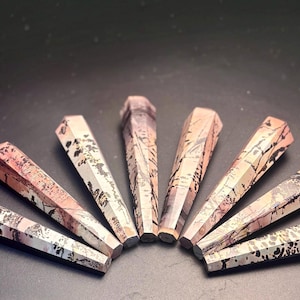 May include: Seven faceted crystal wands arranged in a fan shape. The wands have a geometric design with a mix of colors, including pink, beige, and black. The crystals are displayed on a dark surface.