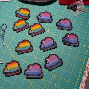 May include: A collection of embroidered patches in the shape of a mouse. The patches feature a rainbow gradient and a bi-pride gradient, each with a black border. The patches are arranged on a green cutting mat.