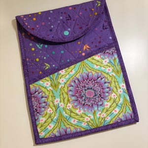 May include: A quilted fabric case with a flap closure. The top section is purple with a star and bird pattern, and the bottom section features a floral design with purple, green, and white colours. A teal button secures the flap.