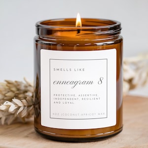 Puede incluir: Una vela encendida en un tarro de vidrio ámbar con una etiqueta blanca. La etiqueta dice "Smells Like enneagram 8" con palabras descriptivas debajo. La vela es de 255 g y está hecha de cera de coco-albaricoque. Una ramita de flores secas está a la izquierda.