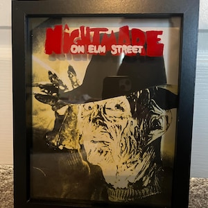 Op de afbeelding: Een ingelijst beeld van Freddy Krueger uit de film "A Nightmare on Elm Street". De afbeelding is in zwart-wit met een gele tint. De titel van de film is in rode letters.