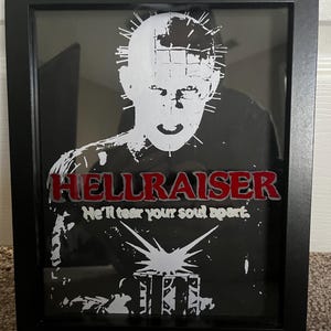 May include: A black framed shadow box with a white silhouette of Pinhead from the movie Hellraiser. The text "Hellraiser" is in red and the text "He'll tear your soul apart." is in white.