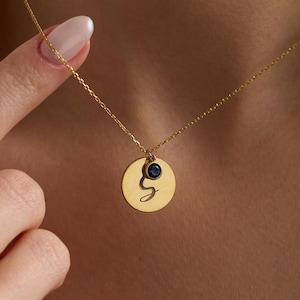 Könnte beinhalten: Eine goldfarbene Halskette mit einem kreisförmigen Anhänger. Der Anhänger hat einen schwarzen Buchstaben "g" und einen kleinen, dunkelblauen Edelstein. Die Halskette wird um den Hals getragen.