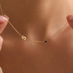 Puede incluir: Un delicado collar de oro con la letra "S" y una pequeña gema azul rectangular. El collar se muestra sobre un fondo de tono de piel, destacando su fina cadena y diseño minimalista. Una elegante pieza de joyería.