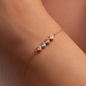 May include: A gold-colored bracelet featuring a delicate chain and a series of square-cut gemstones in pink, blue, yellow, and clear. The bracelet is worn on a person's arm, showcasing the jewelry's design and color palette.