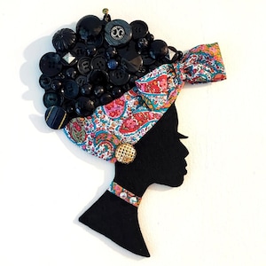 Puede incluir: Silueta negra del perfil de una mujer, adornada con un pañuelo y joyas. El cabello está hecho de varios botones negros. El pañuelo presenta un colorido estampado de cachemira y un botón dorado adorna el cuello.