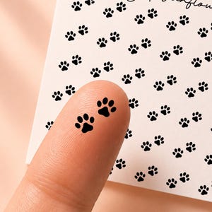 May include: A square white sticker sheet featuring a repeating pattern of small black paw prints. The text "honeyed sunflower" and "CL047" are printed in black at the top. One individual black paw print sticker is applied to a fingertip.
