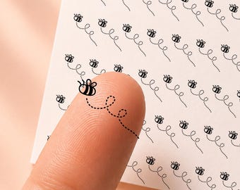 May include: A square white sticker sheet labeled honeyed sunflower CL091 featuring multiple rows of small black line art illustrations of bees with dotted flight paths. One individual bee sticker is applied to a fingertip showing the design.