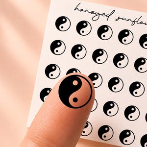 May include: A square sheet of white adhesive stickers featuring a grid of small black and white yin-yang symbols. The text "honeyed sunflower" and "CL100" are printed at the top. One circular yin-yang sticker is applied to a fingertip.