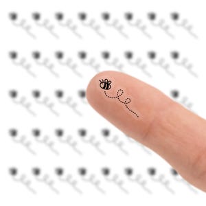 May include: A close-up of a finger with a small bee sticker. The bee is black and yellow with a dotted flight path. The background is white with a pattern of blurred black shapes.