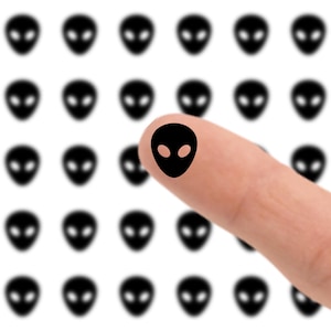 May include: A finger with a black alien head sticker on it, with a background of similar alien head stickers. The alien heads are black with oval eyes, set against a white background.