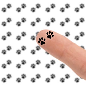 May include: A close-up of a finger with two black paw print stickers. The background features a pattern of blurred black paw prints on a white background. The image is a playful representation of pet-themed designs.