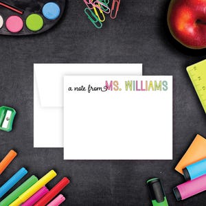 May include: White stationery set with a note card and envelope. The note card reads "a note from MS. WILLIAMS" in black and rainbow-colored text. The image also includes school supplies like markers, a ruler, and an apple.