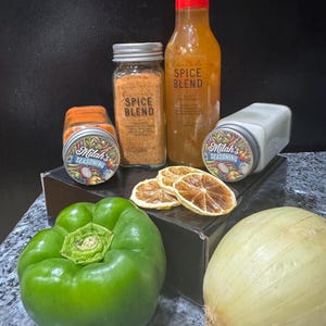 Puede incluir: Una colecci&oacute;n de especias y salsas, incluyendo dos frascos de mezcla de especias, una botella de salsa picante y un frasco de condimento. Los frascos y la botella est&aacute;n etiquetados con el texto "Milah's Seasoning" y "Spice Blend". La imagen tambi&eacute;n incluye un pimiento verde, una cebolla blanca y rodajas de c&iacute;tricos secas.