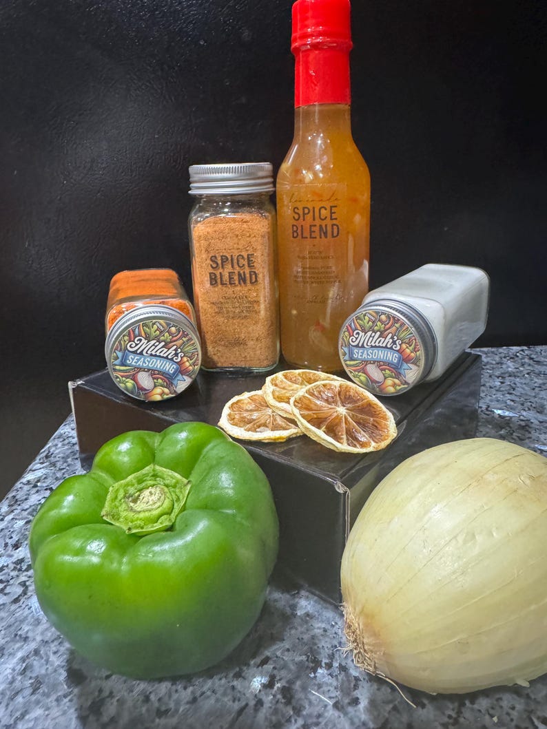 Puede incluir: Una colecci&oacute;n de especias y salsas, incluyendo dos frascos de mezcla de especias, una botella de salsa picante y un frasco de condimento. Los frascos y la botella est&aacute;n etiquetados con el texto "Milah's Seasoning" y "Spice Blend". La imagen tambi&eacute;n incluye un pimiento verde, una cebolla blanca y rodajas de c&iacute;tricos secas.