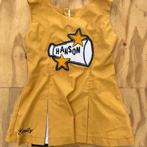May include: A mustard yellow sleeveless dress featuring a white megaphone applique with the text HANSON and two orange stars. The dress has a round neckline, a back zipper, and two front pleats with white fabric inserts. The name Emily is embroidered in black on the lower left side.