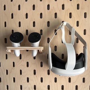 Könnte beinhalten: Ein VR-Headset und Controller werden auf einer Holz-Lochwand präsentiert. Das Headset ist weiß und schwarz, mit verstellbaren Riemen. Zwei weiße Controller mit schwarzen Oberteilen ruhen auf einem kleinen Holzregal. Die Lochwand hat gleichmäßig verteilte ovale Löcher.