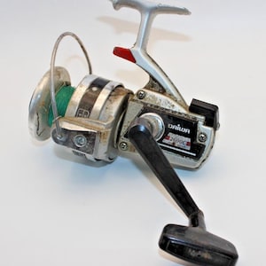 May include: A silver and black Daiwa 2600C spinning fishing reel with a green fishing line wound on the spool. The reel features a metal body, a black plastic handle, a silver bail arm, and a red accent near the top of the reel housing.
