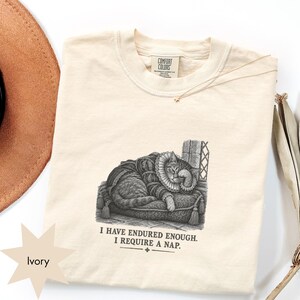 May include: An ivory t-shirt featuring a black and white graphic of a cat wearing a ruff collar, resting on a tasseled cushion. The text below the cat reads, "I HAVE ENDURED ENOUGH. I REQUIRE A NAP." The shirt has a Comfort Colors brand label.