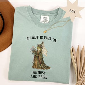 May include: A light green t-shirt with the text "M'LADY IS FULL OF WHIMSY AND RAGE" and a graphic of a frog in a dress holding flowers and a flag. A gold necklace is draped across the shirt.