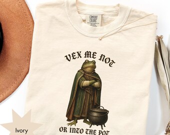 May include: An ivory t-shirt featuring a graphic of a frog wearing a green cloak and standing next to a black cauldron. The text above the frog reads VEX ME NOT and the text below reads OR INTO THE POT. The shirt has a Comfort Colors brand label.
