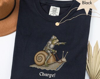 May include: A black t-shirt featuring a graphic of a frog wearing medieval armor and riding a snail. The frog is pointing forward with one hand, and the word "Charge!" is printed in white text below the illustration. The shirt has a Comfort Colors label.