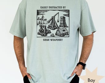 May include: A light sage green short-sleeve t-shirt featuring a black ink-style graphic of medieval siege weaponry, including a trebuchet, a battering ram, and a castle. The text reads "EASILY DISTRACTED BY" above and "SIEGE WEAPONRY" below.
