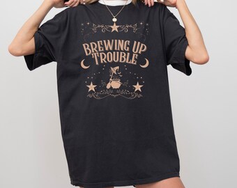May include: Black t-shirt with the phrase "Brewing Up Trouble" in a tan, vintage-style font. The design includes stars, moons, and a witch's cauldron. A casual, oversized fit.