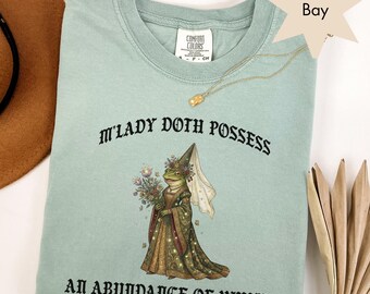May include: A light green t-shirt with the text "M'LADY DOTH POSSESS AN ABUNDANCE OF WHIMSY" and a graphic of a frog in a medieval dress. A gold necklace is draped across the shirt. The shirt is made of a soft material.