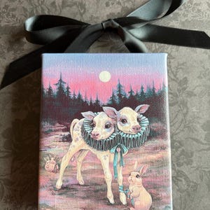May include: A small canvas art print featuring a whimsical scene with two-headed calves wearing ruffled collars, a rabbit, and a forest backdrop under a pink and blue sky. A black ribbon is tied at the top.