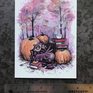 May include: A watercolor illustration of an owl wearing a scarf and reading a book in a forest setting. The owl is surrounded by pumpkins and a cup of tea. The background is a soft pink and purple.