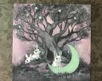 May include: A watercolor painting of three white rabbits under a large tree with a crescent moon. The moon is painted in a light green color and the background is a soft purple.