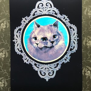 May include: A painting of a gray cat with two sets of eyes, framed in a silver ornate frame against a dark blue background.