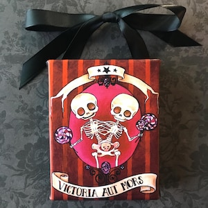 May include: A small canvas painting with a red, white, and black color scheme. The painting features two skeletons holding lollipops. The skeletons are conjoined at the hips. The painting has the text "VICTORIA AUT MORS" at the bottom.