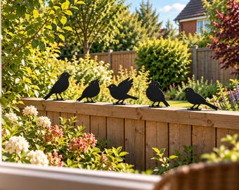 May include: A set of five black metal bird silhouettes arranged in a row along the top of a wooden fence. Each bird figure features a distinct pose, including standing, perched, and with wings slightly spread, creating a decorative garden display.