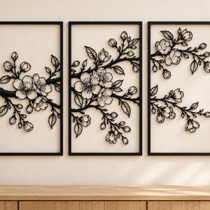 May include: A triptych wall art set featuring three rectangular black frames. Each frame contains a black metal cutout of cherry blossom branches with detailed flowers, buds, and leaves, creating a continuous floral design across the panels.