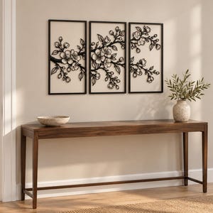 May include: A set of three rectangular wall art panels featuring black metal cherry blossom branches against a light background, displayed above a long, dark wooden console table with a stone bowl and a vase with green leaves.