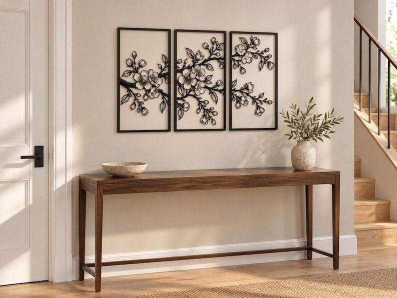 May include: A set of three rectangular wall art panels featuring black metal cherry blossom branches against a light background, displayed above a long, dark wooden console table with a stone bowl and a vase with green leaves.
