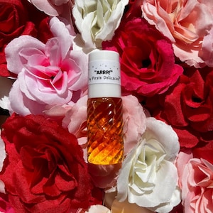 May include: A small, clear glass bottle with a white label that reads "ARRR!" by Pirate Delicacies. The bottle is filled with a golden liquid and is surrounded by pink and red roses.