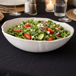 May include: A large, off-white ceramic bowl filled with a fresh salad. The salad includes green lettuce, red cherry tomatoes, red onion slices, cucumber, and crumbled white cheese. The bowl sits on a black tablecloth, with wine glasses and candles in the background.